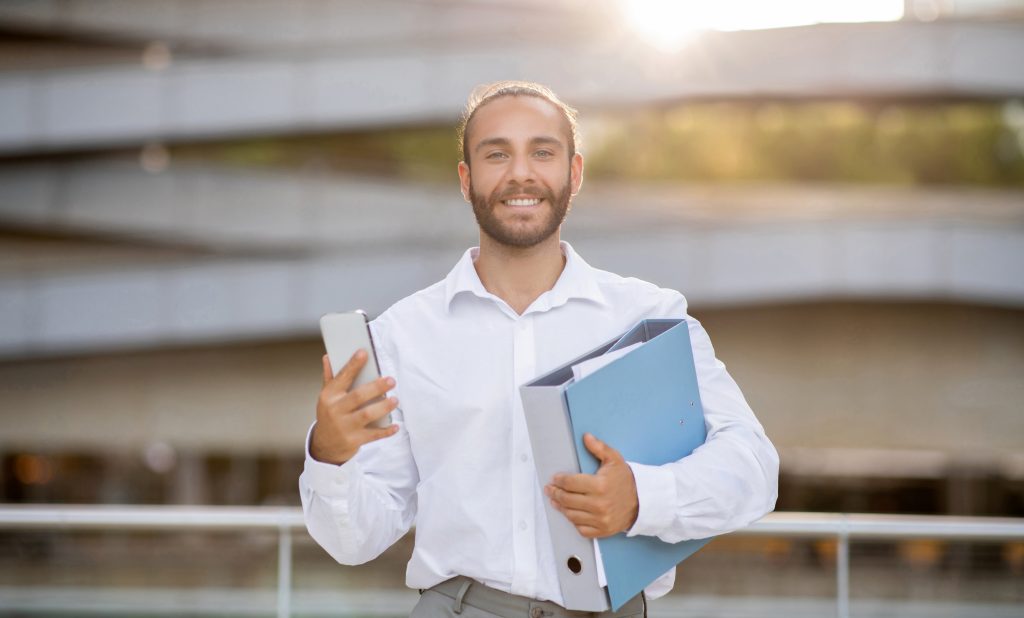 Young professional man standing outdoors with smartphone and documents folder in hands