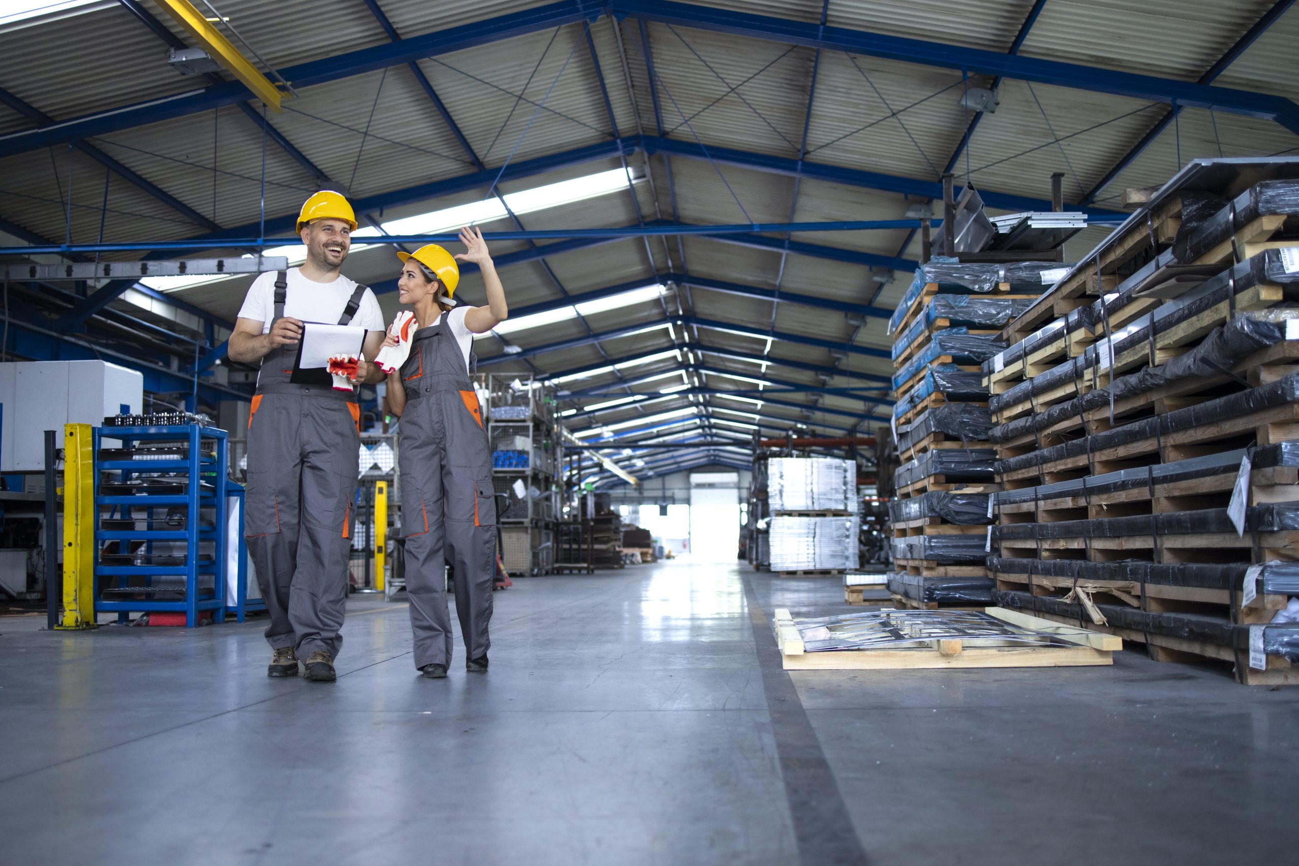 Factory workers in work wear and yellow helmets walking through industrial production hall and discussing about organization.